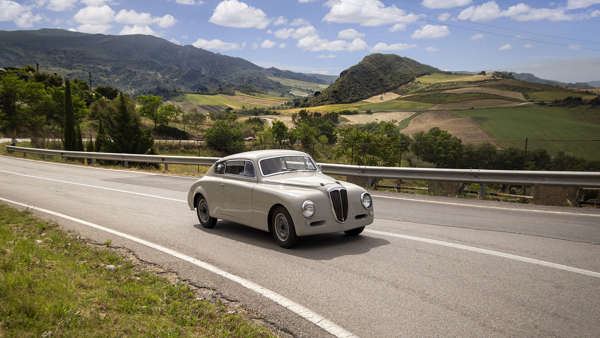Klassischer Oldtimer fährt auf Landstraße durch hügelige Landschaft mit Feldern, Bergen und blauem Himmel
