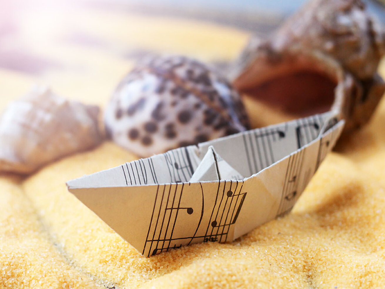 © sabdiz - AdobeStock.com Gefaltetes Papierschiffchen auf dekorativem Sand mit Muscheln und maritimer Tischdekoration.
