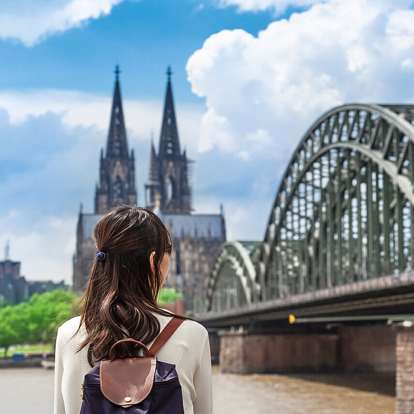 Frau mit Rucksack blickt auf den Kölner Dom und die Hohenzollernbrücke am Rhein