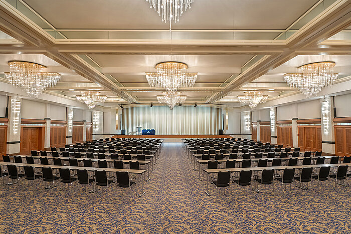 Fürst-von-Metternich-Saal mit großer Bühne, Reihenbestuhlung, eleganten Kronleuchtern und hellem Ambiente.