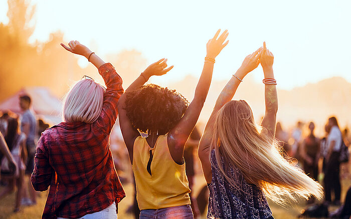 Drei Freunde tanzen mit erhobenen Armen im Sonnenuntergang bei einem Musikfestival im Freien.