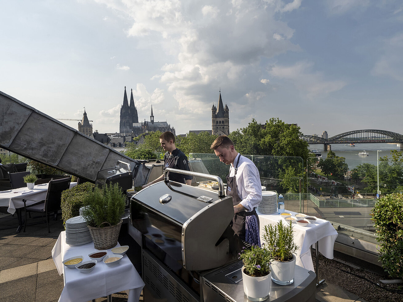 Dachterrasse des Maritim Hotels Köln mit Blick auf den Dom und die Hohenzollernbrücke. Köche bereiten ein BBQ vor.