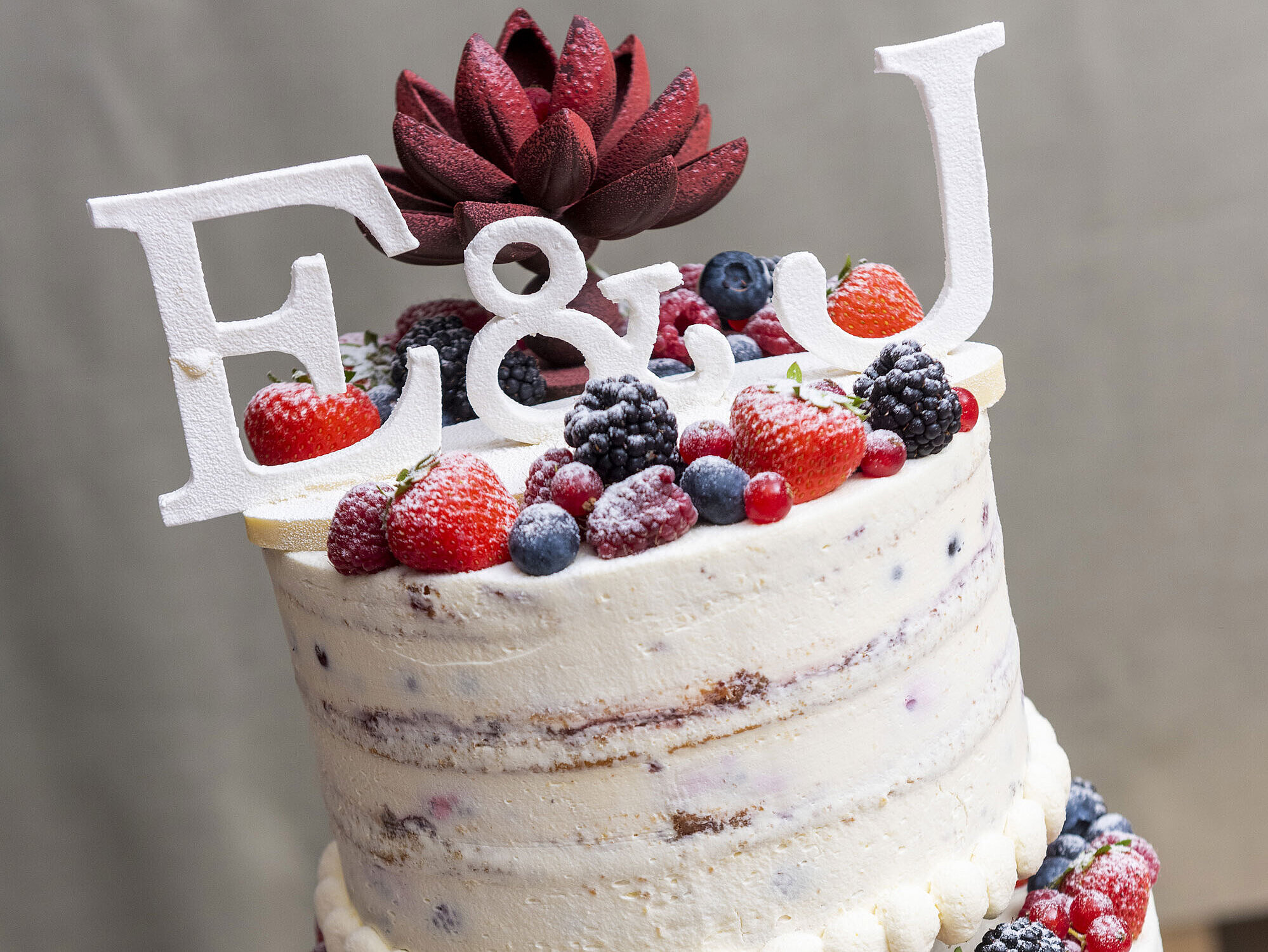 Hochzeitstorte mit Beeren, dekoriert mit einem "E&J" Topper und einer roten Blume, perfekter Genuss für besondere Momente.
