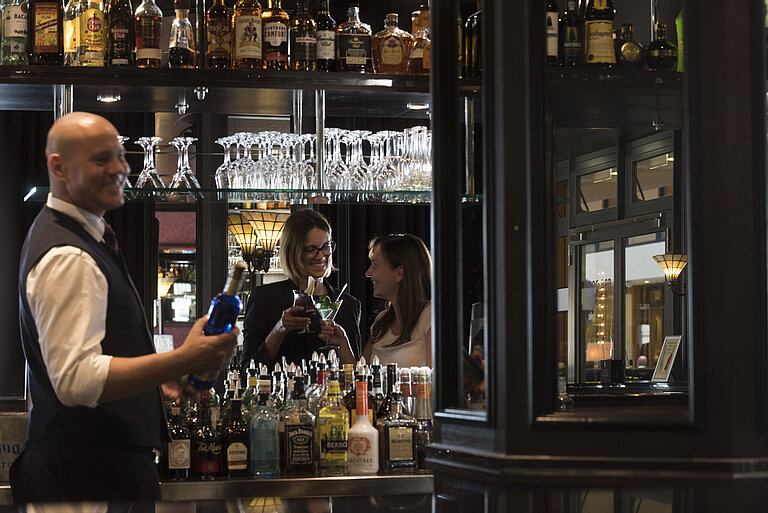 Fröhliche Gäste genießen Cocktails an der Bar des Maritim Hotel Dresden, serviert von einem freundlichen Barkeeper.