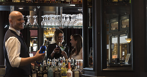 Fröhliche Gäste genießen Cocktails an der Bar des Maritim Hotel Dresden, serviert von einem freundlichen Barkeeper.