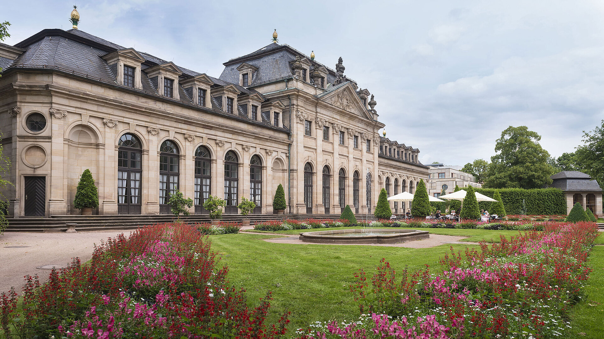 Die historische Orangerie in Fulda mit barocker Fassade, Treppe und blühendem Garten im Vordergrund.