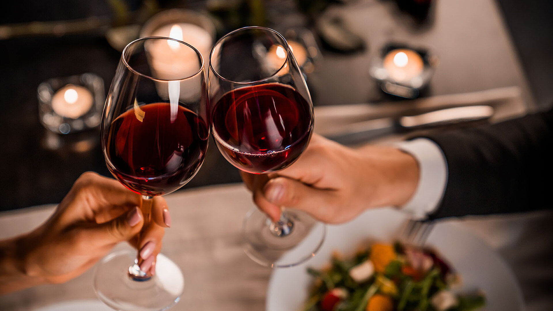 © InsideCreativeHouse - AdobeStock.com Zwei Gläser Rotwein stoßen bei einem romantischen Dinner an