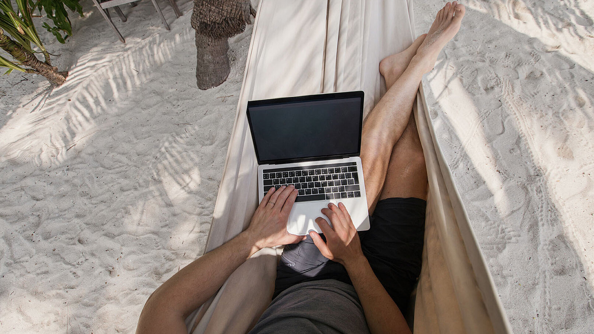 Person arbeitet entspannt mit Laptop in einer Hängematte am Strand unter Palmen.