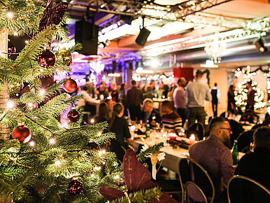 © bilderstöckchen - AdobeStock.com Weihnachtsfeier mit festlich geschmücktem Baum und Gästen im festlichen Saal