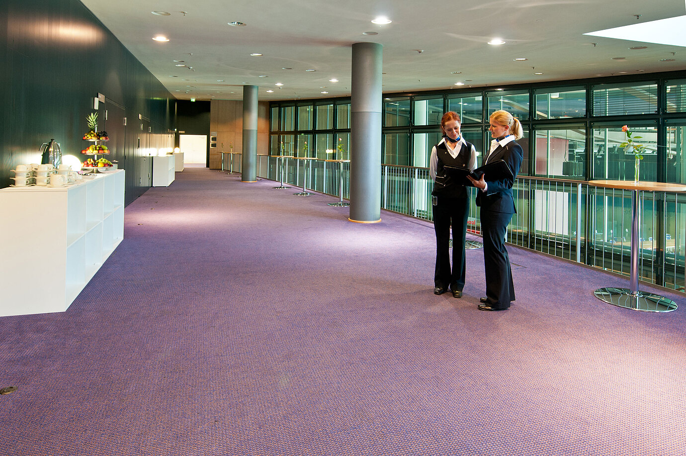  Foyer im Maritim Hotel Dresden mit zwei Mitarbeitenden in Uniform, Stehtischen und einer Obststation.