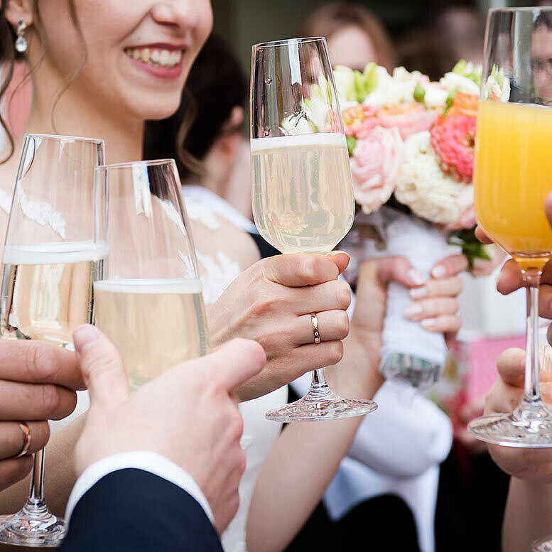 © ilfotokunst - AdobeStock.com Hochzeitsgäste stoßen mit Sekt und Orangensaft an, während die Braut einen Blumenstrauß hält