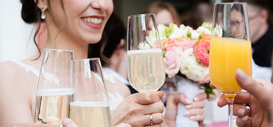 Hochzeitsgäste stoßen mit Sekt und Orangensaft an, während die Braut einen Blumenstrauß hält