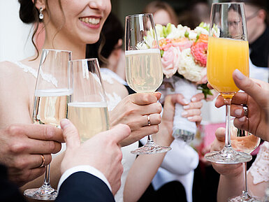 © ilfotokunst - AdobeStock.com Hochzeitsgäste stoßen mit Sekt und Orangensaft an, während die Braut einen Blumenstrauß hält