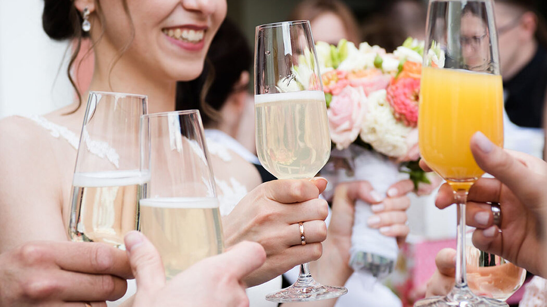 © ilfotokunst - AdobeStock.com Hochzeitsgäste stoßen mit Sekt und Orangensaft an, während die Braut einen Blumenstrauß hält