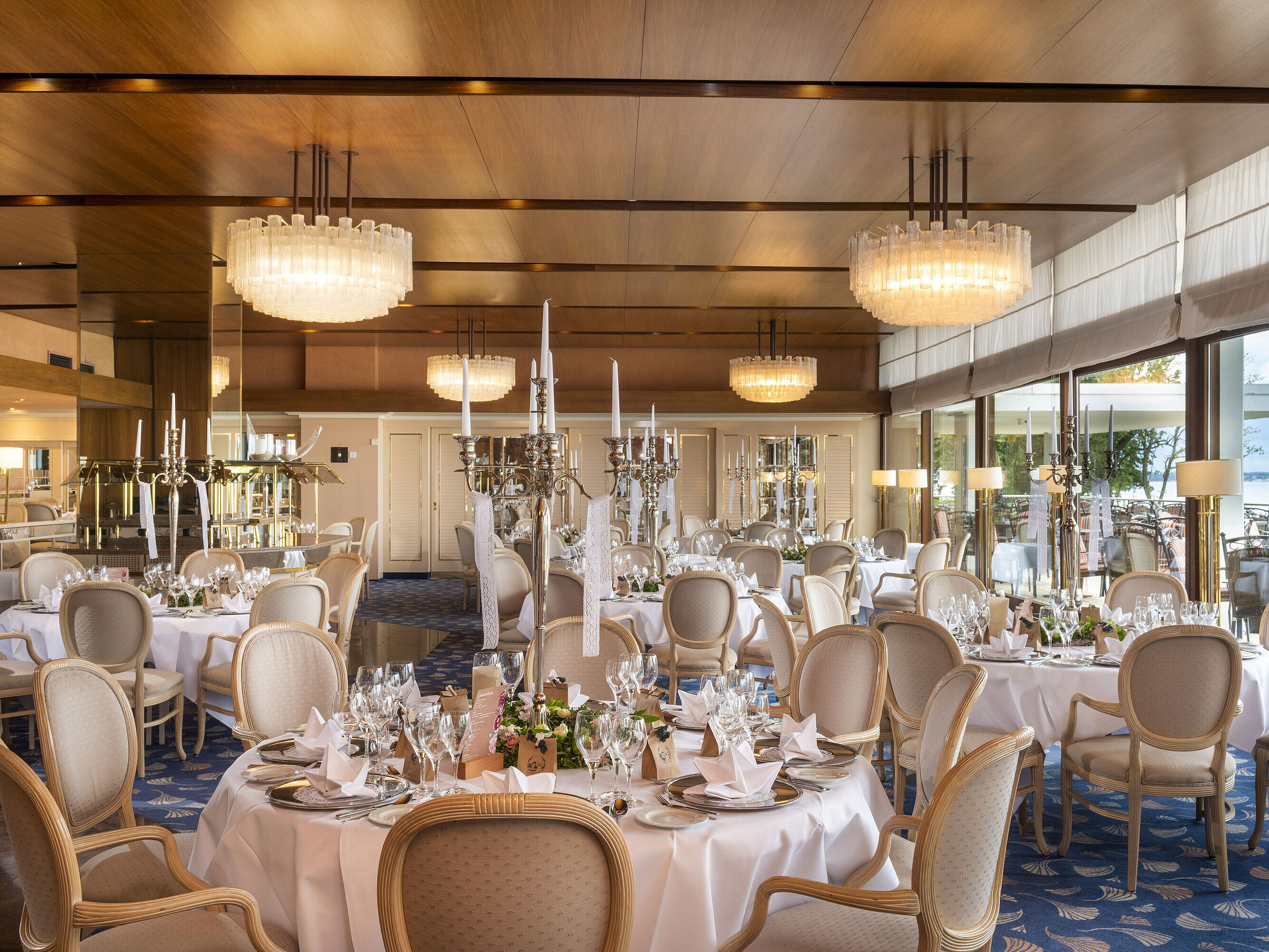 Elegantes Restaurant mit runden Tischen, Kronleuchtern und Panoramafenstern, die einen Ausblick auf die Terrasse bieten.
