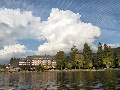 Außenansicht des Maritim Hotel Titisee mit Bäumen und Seeufer bei blauem Himmel und weißen Wolken
