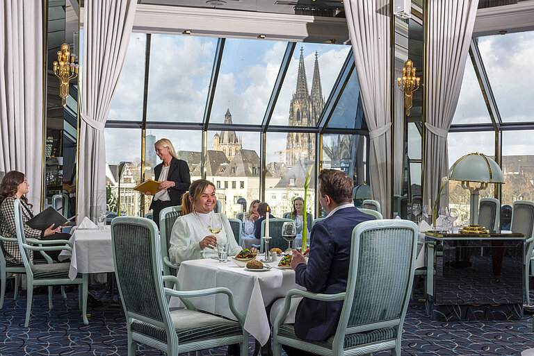 Restaurant "Bellevue" im Maritim Hotel Köln mit Gästen, die speisen, und Blick auf den Kölner Dom durch große Fenster.