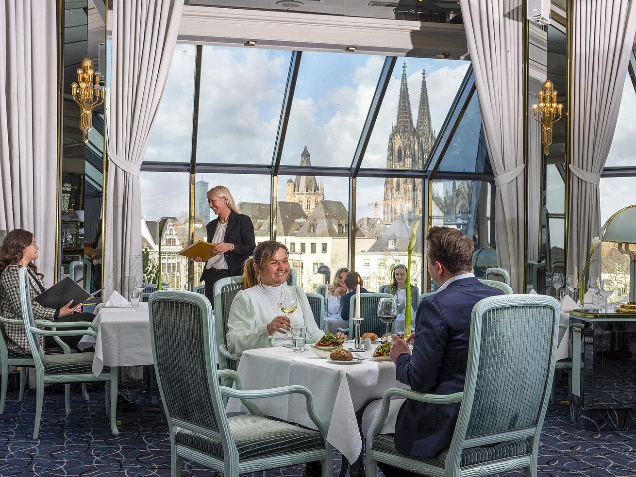 Restaurant "Bellevue" im Maritim Hotel Köln mit Gästen, die speisen, und Blick auf den Kölner Dom durch große Fenster.