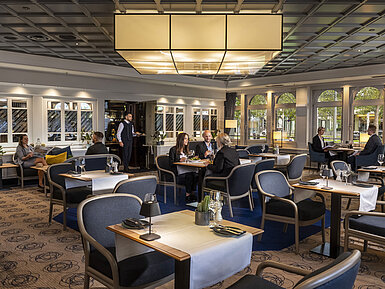 Elegantes Restaurant mit Gästen, die speisen und entspannen. Moderne Einrichtung und große Fenster mit Ausblick.