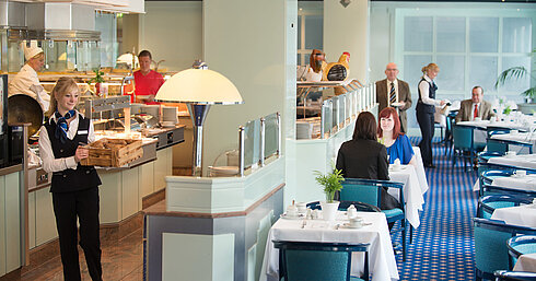 Eine Kellnerin bringt frische Brötchen zum Frühstücksbereich im Maritim Hotel Magdeburg, während Gäste am Tisch sitzen.