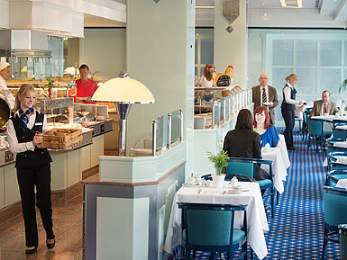 Eine Kellnerin bringt frische Brötchen zum Frühstücksbereich im Maritim Hotel Magdeburg, während Gäste am Tisch sitzen.