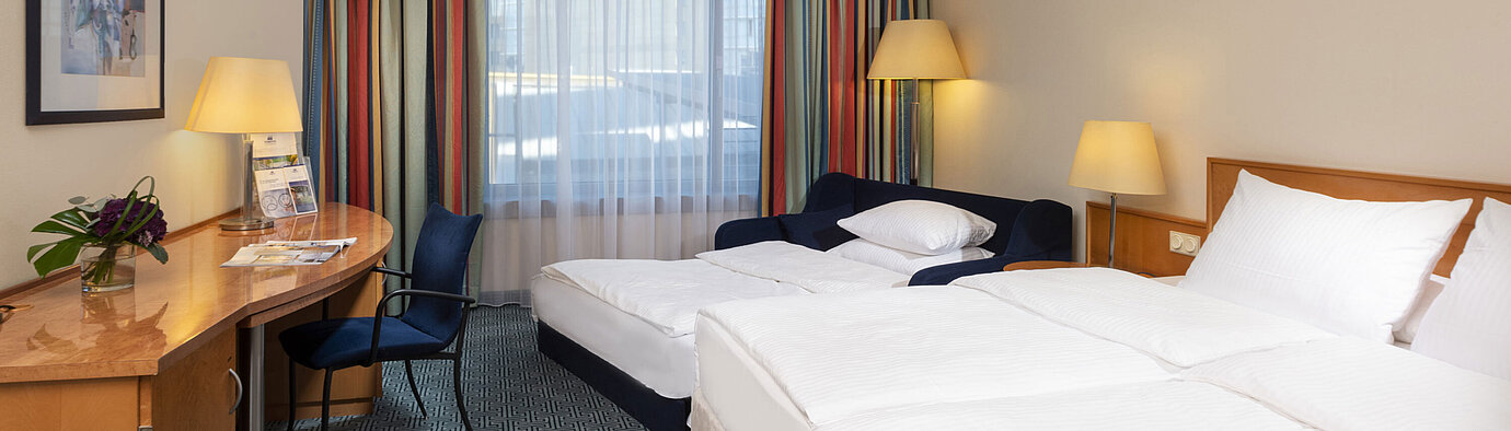 Classic Familienzimmer Classic Familienzimmer im Maritim Hotel Frankfurt mit Doppelbett, Schlafsofa, Schreibtisch und bunten Vorhängen.