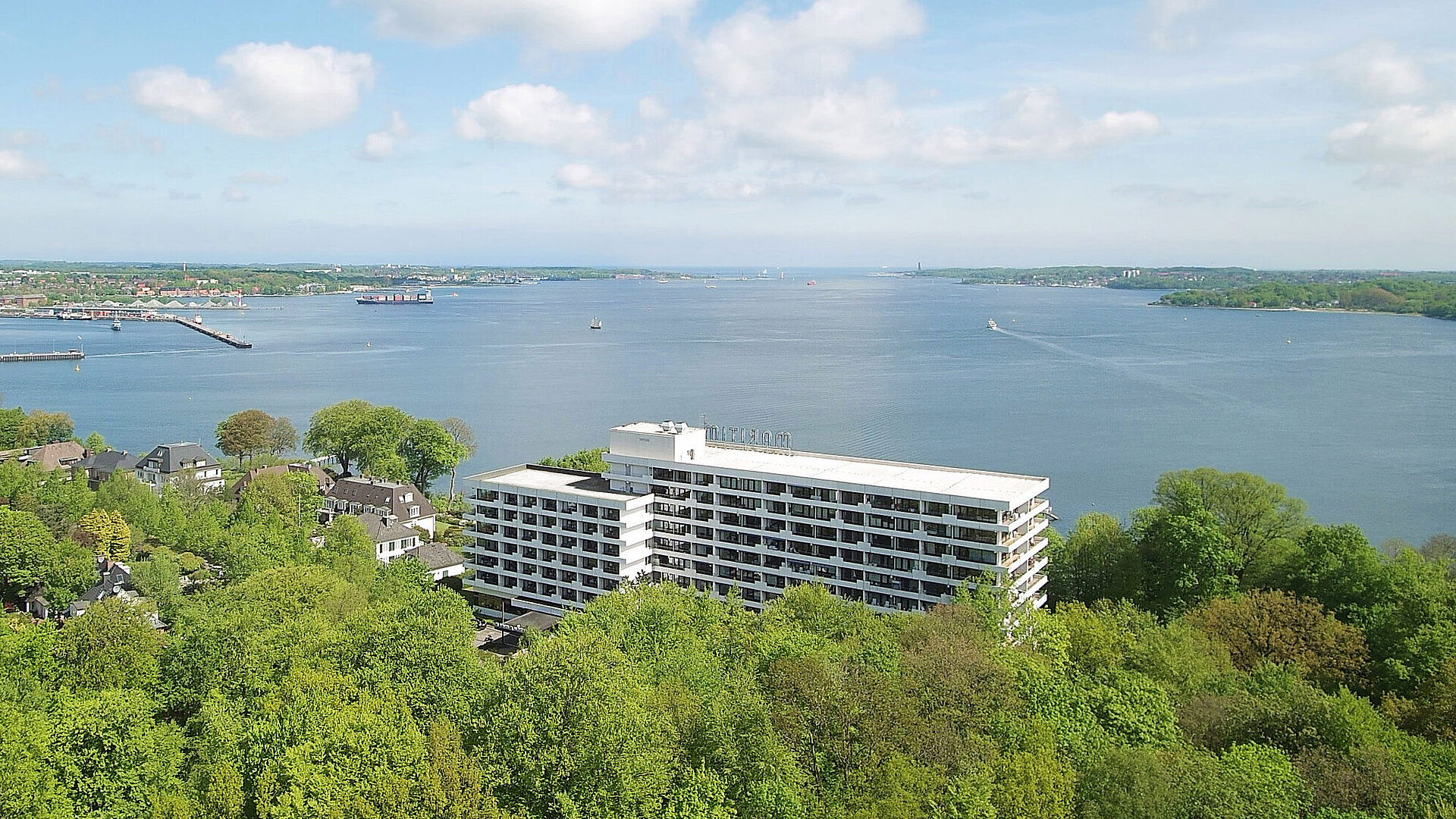 Luftaufnahme des Maritim Hotels Kiel, umgeben von grüner Landschaft und der Kieler Förde, bei strahlend blauem Himmel.