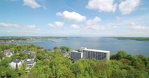 Luftaufnahme des Maritim Hotels Kiel, umgeben von grüner Landschaft und der Kieler Förde, bei strahlend blauem Himmel.