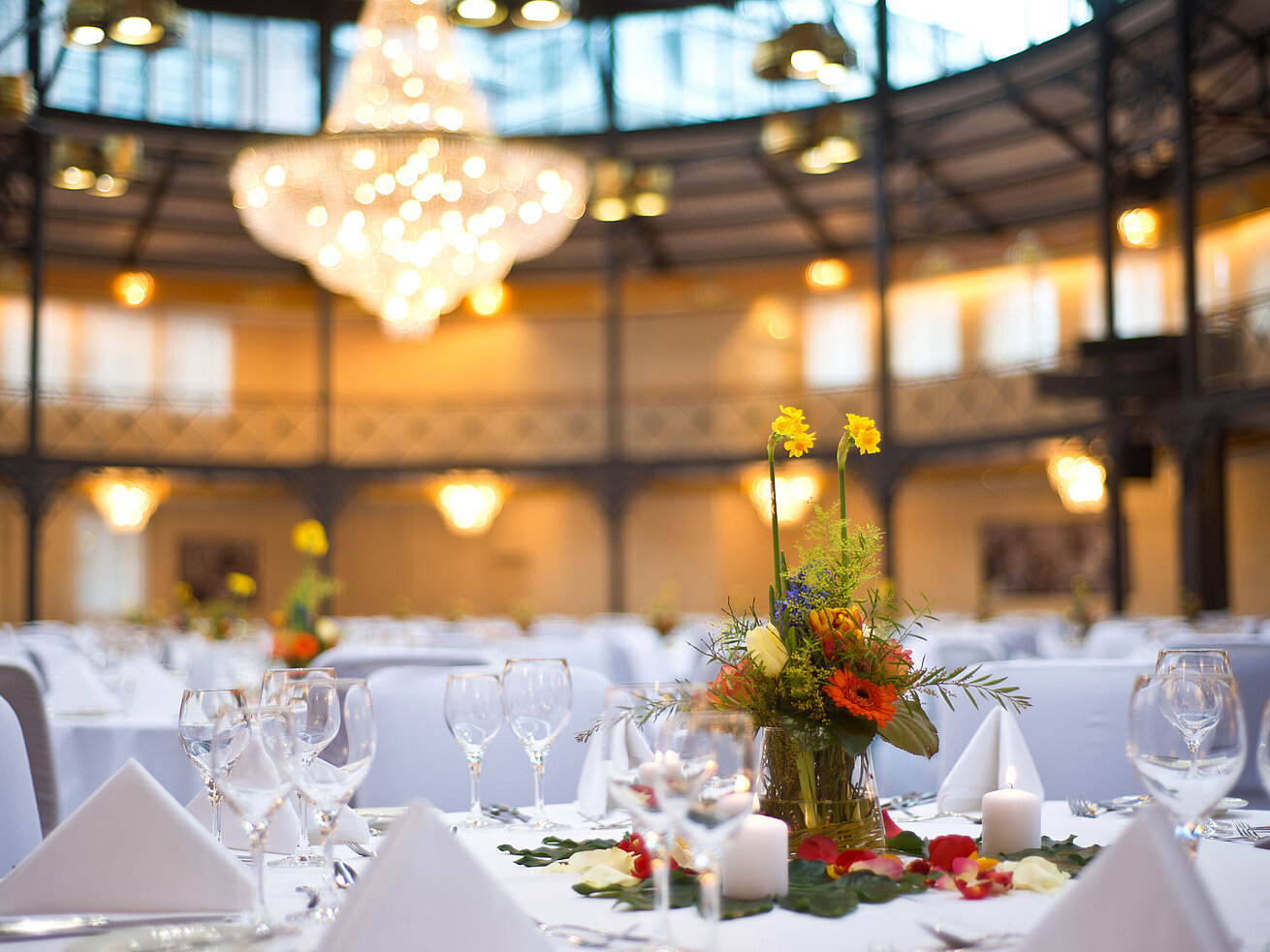 Nahaufnahme einer Tischdekoration mit Blumen und Kerzen, im Hintergrund ein großer Kronleuchter