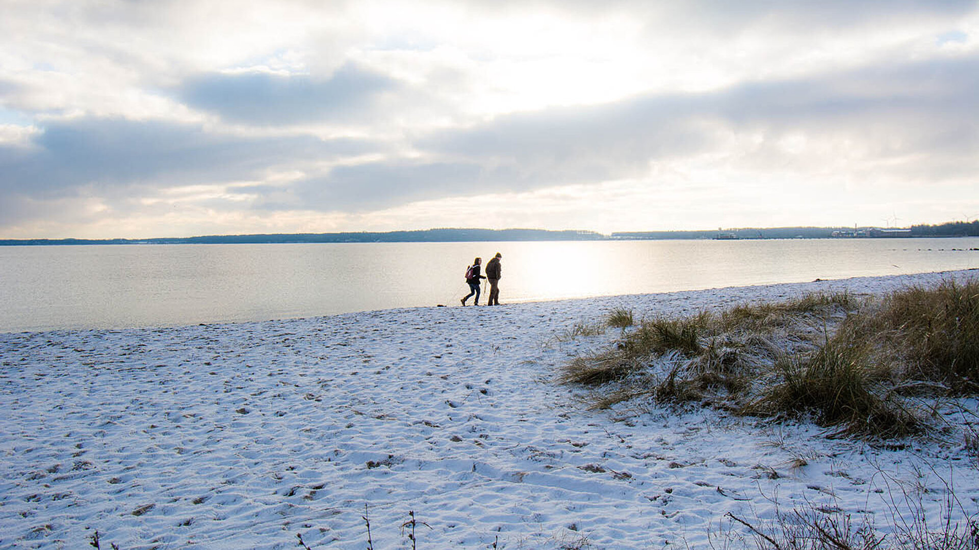 Verschneiter Ostseestrand mit zwei Spaziergängern am Wasser unter winterlichem Himmel