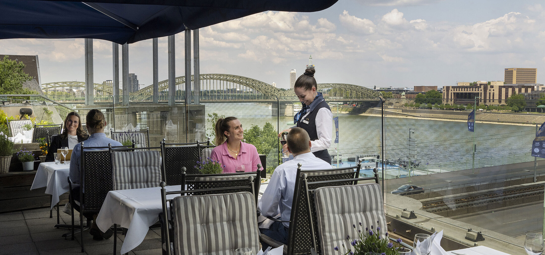 Gäste genießen den Service auf der Dachterrasse des Maritim Hotels Köln mit Blick auf den Rhein und die Hohenzollernbrücke.