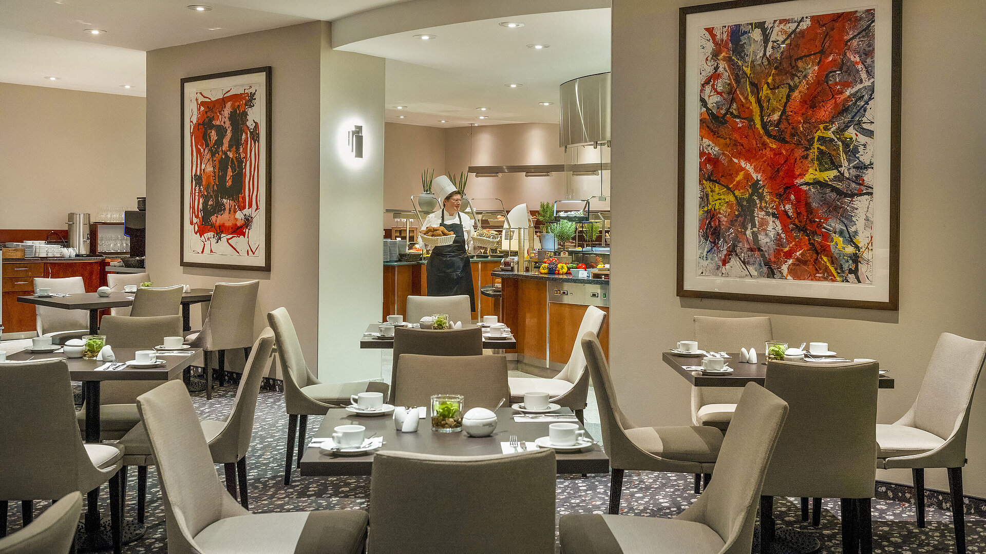 Restaurant im Maritim proArte Hotel Berlin mit moderner Kunst an den Wänden, gedeckten Tischen und einem Koch am Buffet.