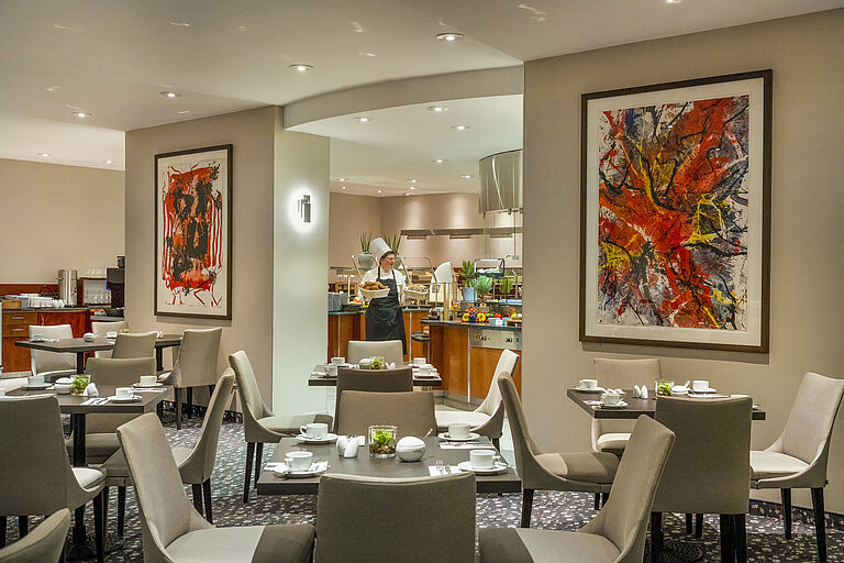 Restaurant im Maritim proArte Hotel Berlin mit moderner Kunst an den Wänden, gedeckten Tischen und einem Koch am Buffet.