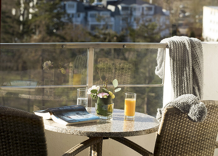 Balkontisch mit Getränken, Buch und Blumen im Maritim Seehotel Timmendorfer Strand mit Aussicht auf Bäume und Gebäude.