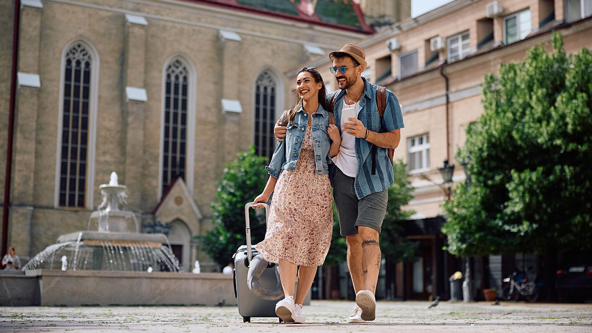 Junges Paar mit Rollkoffer auf Städtereise vor historischer Kirche mit Brunnen bei Sonnenschein
