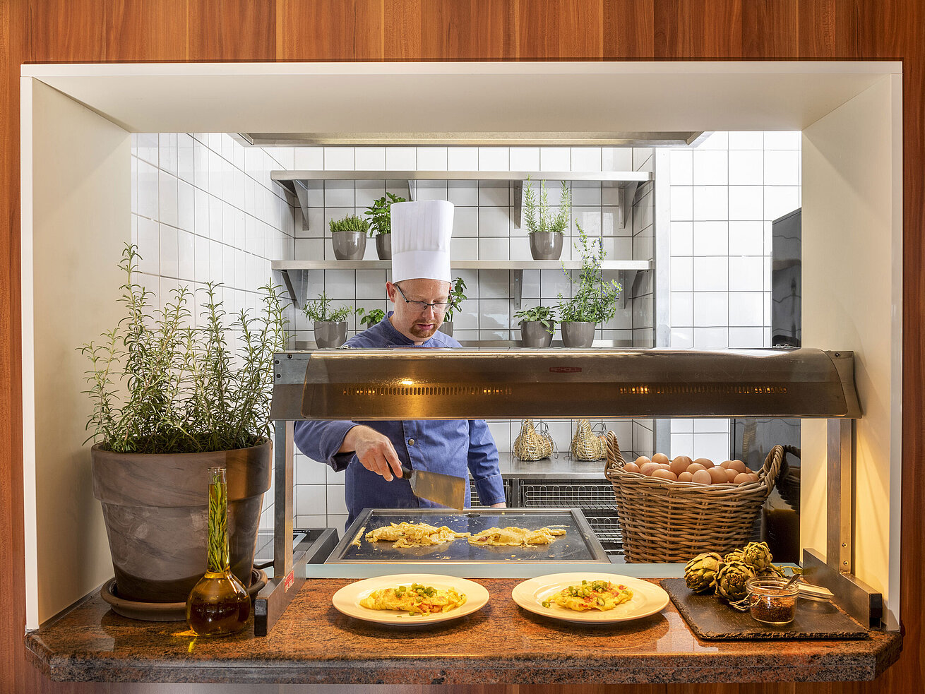 Ein Koch bereitet Omeletts an einer Bratstation zu, umgeben von frischen Kräutern. Zwei Teller mit Essen stehen bereit.