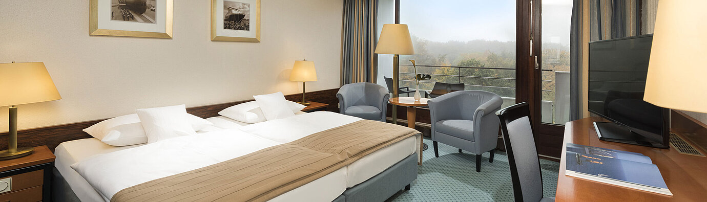 Classic Zimmer im Maritim Hotel Kiel mit großem Doppelbett, Sitzbereich, Schreibtisch und Balkon mit Blick ins Grüne.