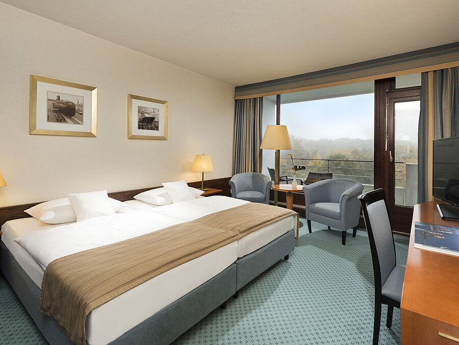 Classic Zimmer im Maritim Hotel Kiel mit großem Doppelbett, Sitzbereich, Schreibtisch und Balkon mit Blick ins Grüne.