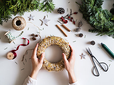 Adventskranz gestalten © Halfpoint - AdobeStock.com Hände gestalten einen Adventskranz auf einem Tisch mit Tannenzweigen, Schleifen und Bastelmaterial.