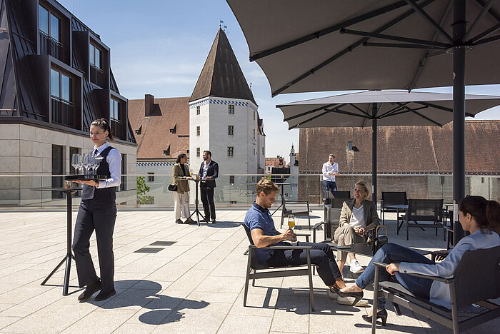 Gäste genießen Getränke auf der Dachterrasse des Maritim Hotel Ingolstadt mit Blick auf die Altstadt.