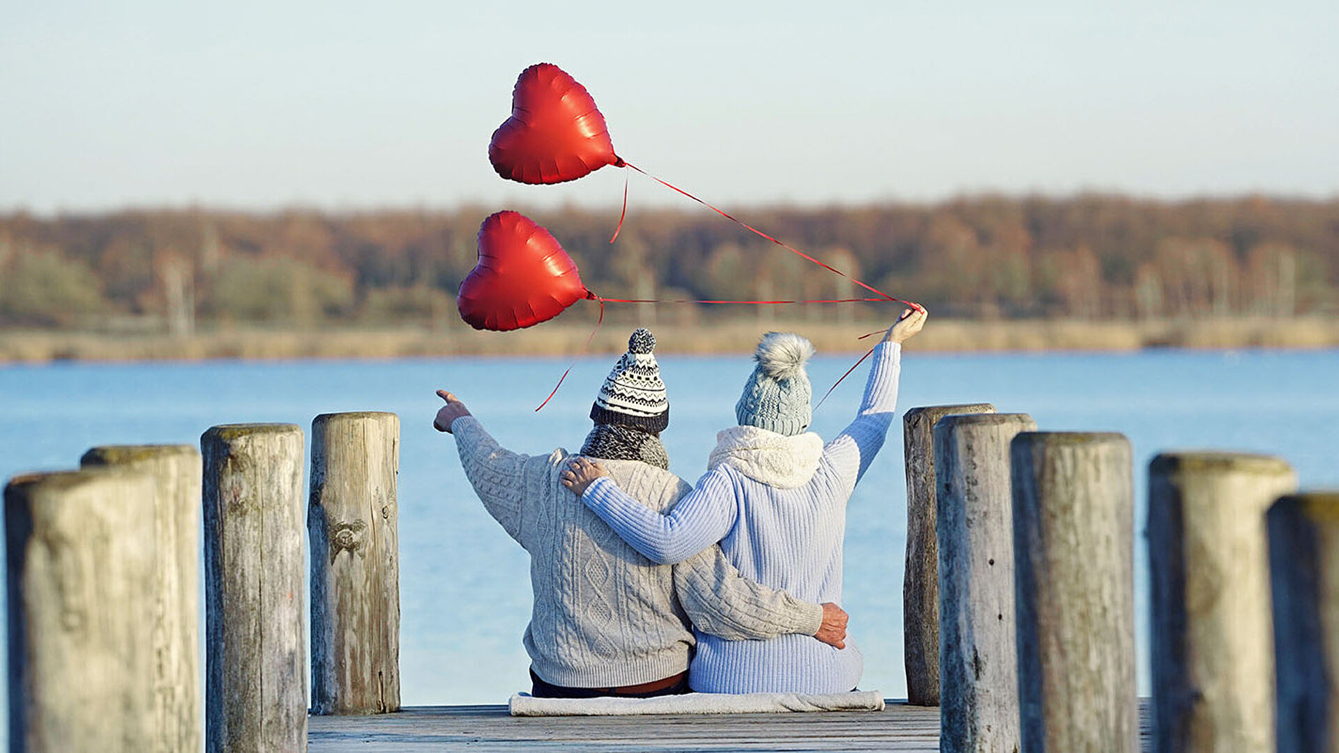 © Jenny Sturm - AdobeStock.com Paar wirft Herzluftballons auf Steg am See in die Luft – romantischer Moment am Wasser