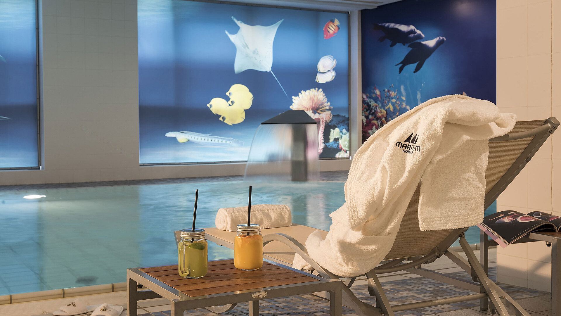Schwimmbad Entspannungsbereich am Pool im Maritim proArte Berlin mit Liegestuhl, Handtuch und frischen Säften neben einem Wasserfall.