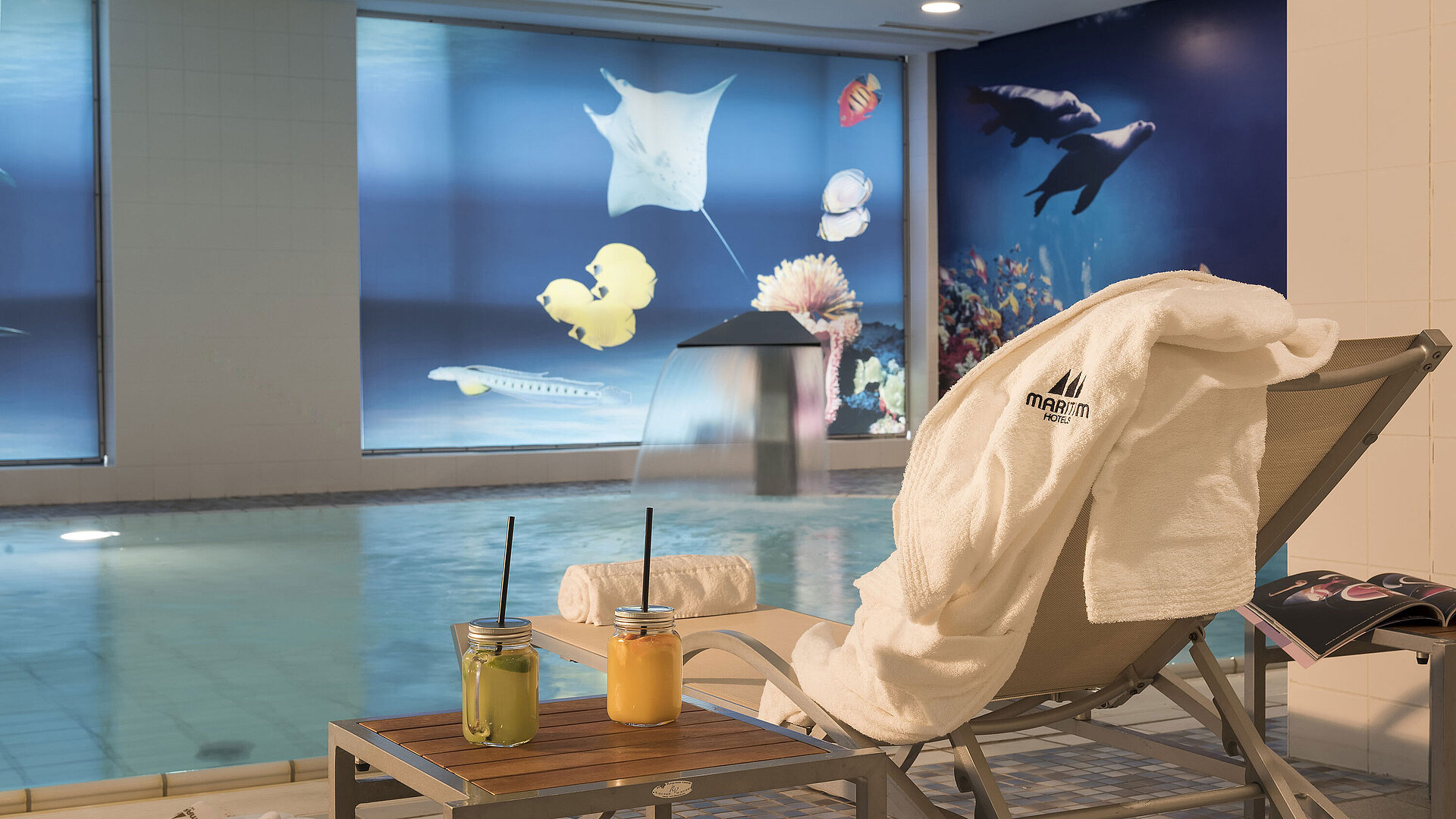 Schwimmbad Entspannungsbereich am Pool im Maritim proArte Berlin mit Liegestuhl, Handtuch und frischen Säften neben einem Wasserfall.