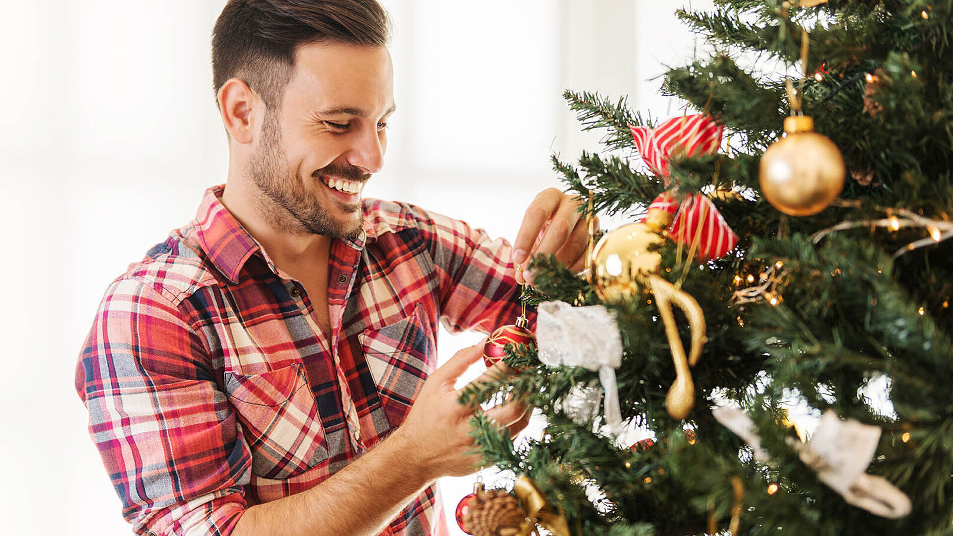 Person schmückt einen Weihnachtsbaum mit goldenen und roten Ornamenten und wirkt dabei fröhlich und entspannt.