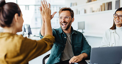 Zwei Kollegen im Büro geben sich lachend ein High Five als Zeichen erfolgreicher Teamarbeit.