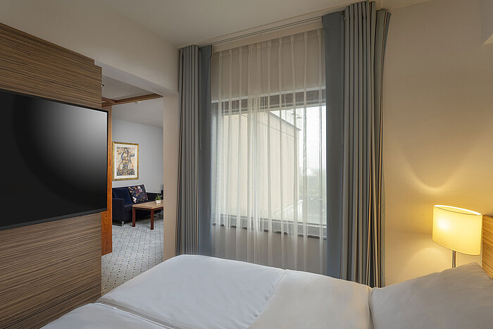 Junior Suite im Maritim Darmstadt mit gemütlichem Bett, separatem Wohnbereich und bodentiefem Fenster für viel Tageslicht.