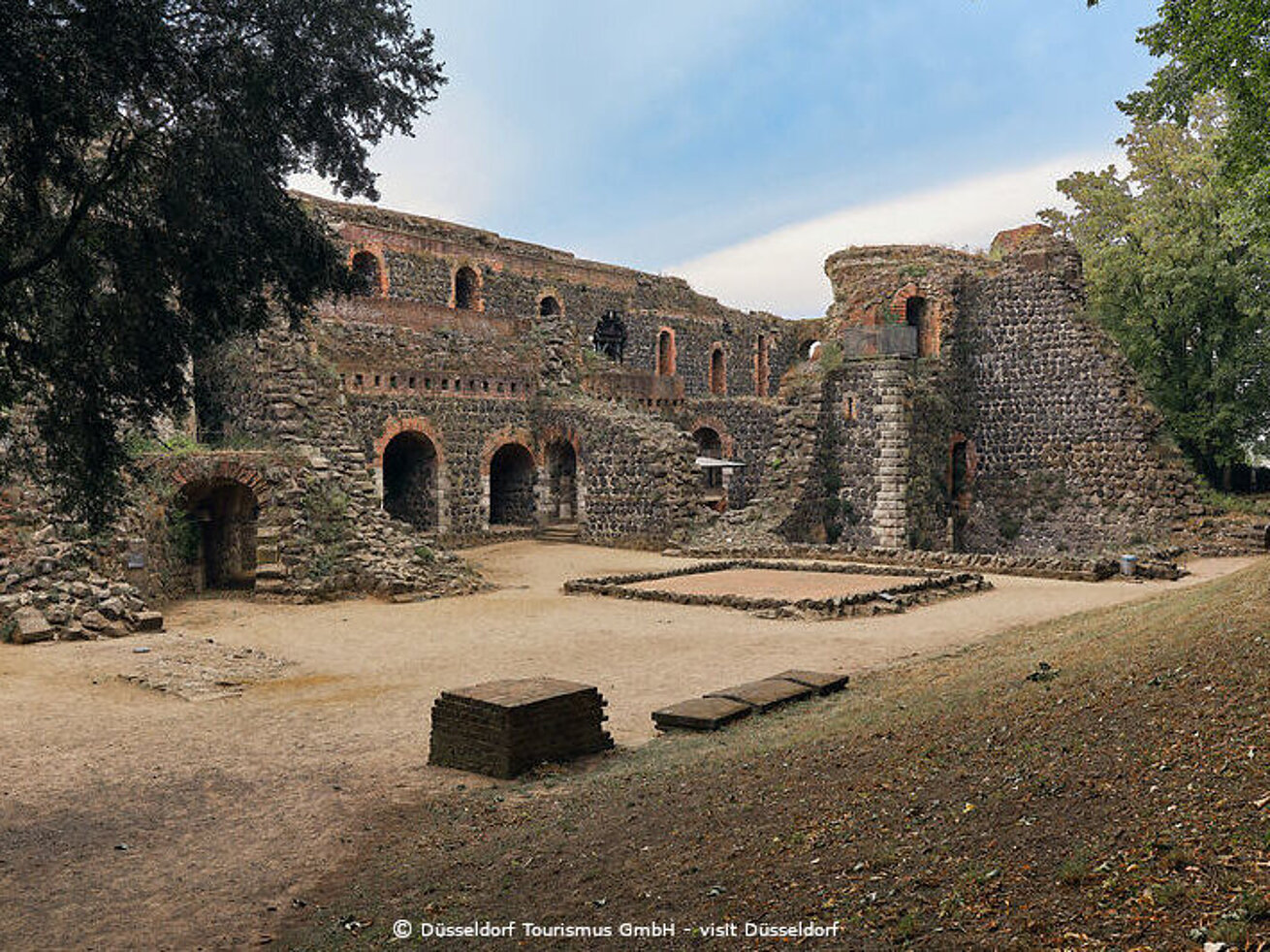 Historische Burgruine aus Naturstein mit Innenhof und Bäumen in sonniger Landschaft