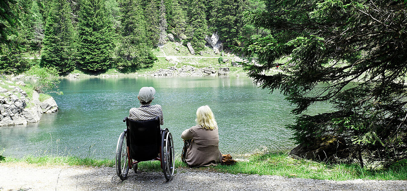 Zwei Personen, eine im Rollstuhl, sitzen am Seeufer und blicken auf den Wald