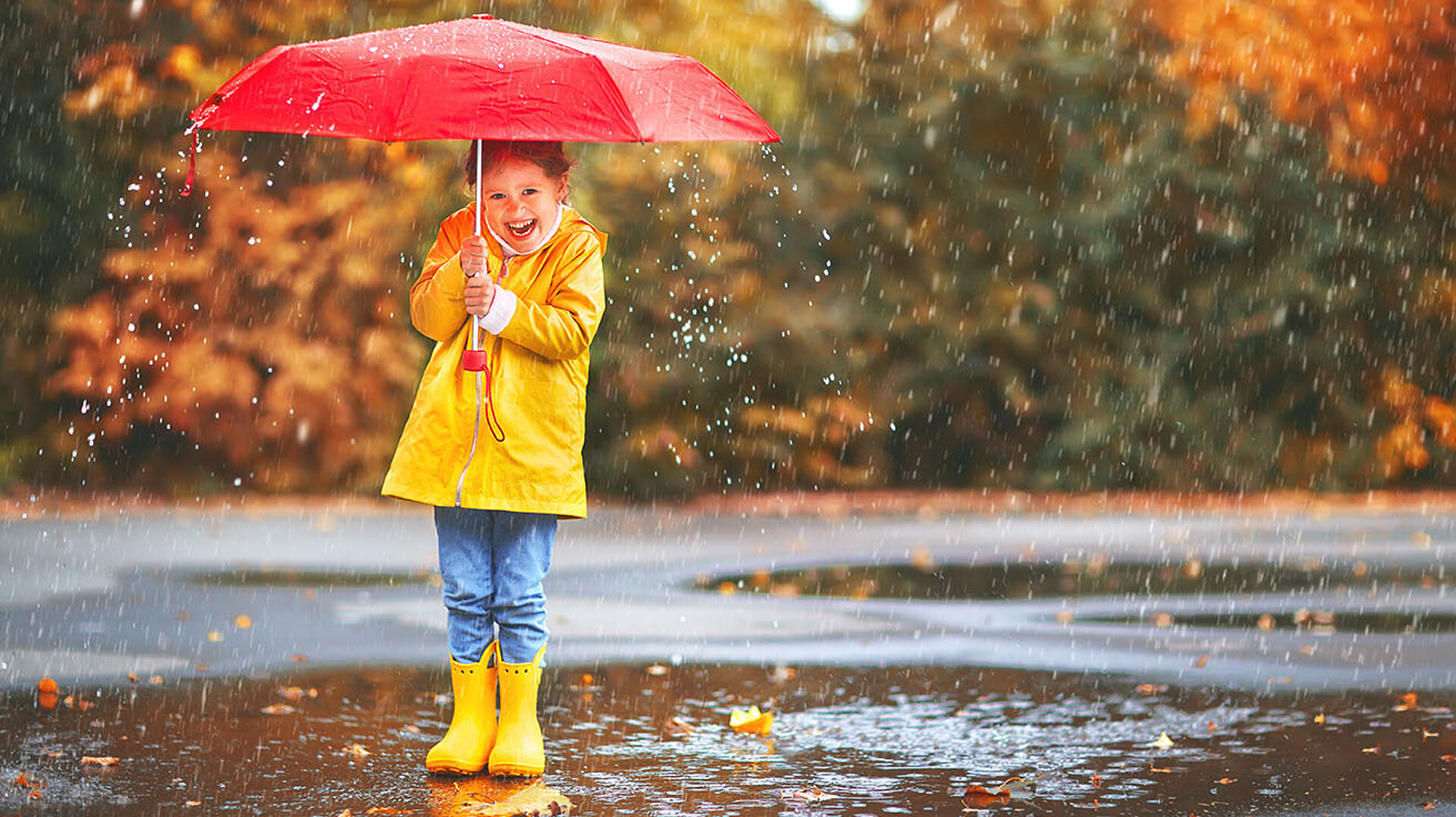 Kind mit gelbem Regenmantel und Stiefeln hält roten Schirm und lacht im herbstlichen Regen.