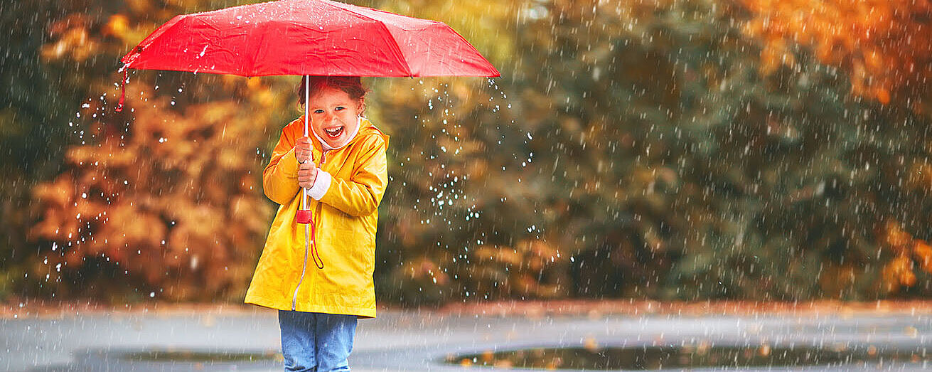 © JenkoAtaman Kind mit gelbem Regenmantel und Stiefeln hält roten Schirm und lacht im herbstlichen Regen.